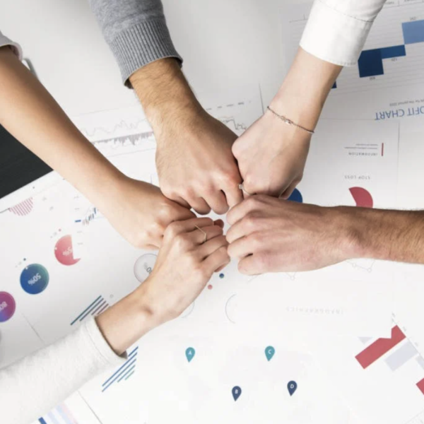 Five people fist bump each other over a desk with data papers on it