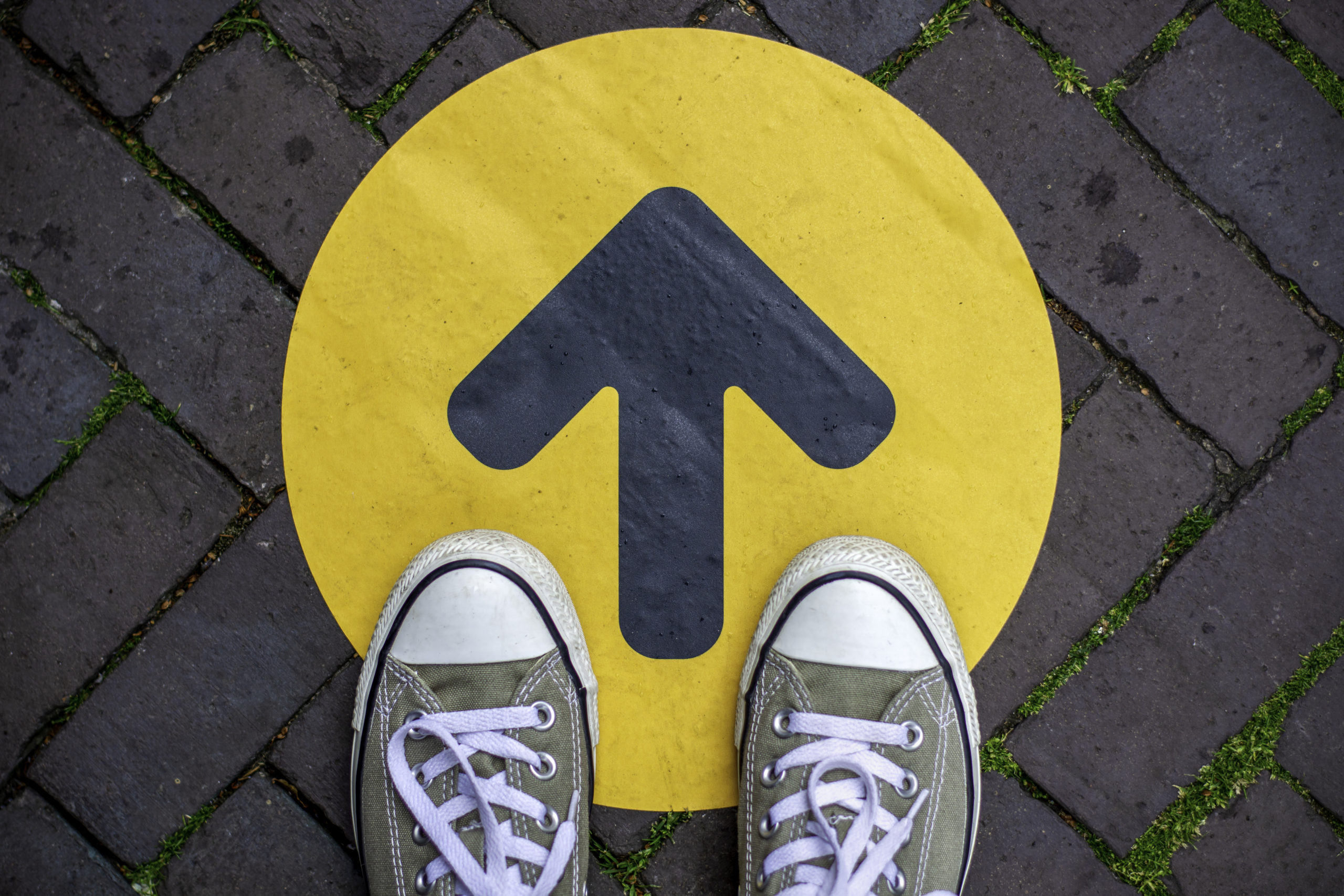 Feet wearing sneakers in front of arrow on road outdoors. Direction arrow in front of a store for the social distancing during the covid-19 pandemic.