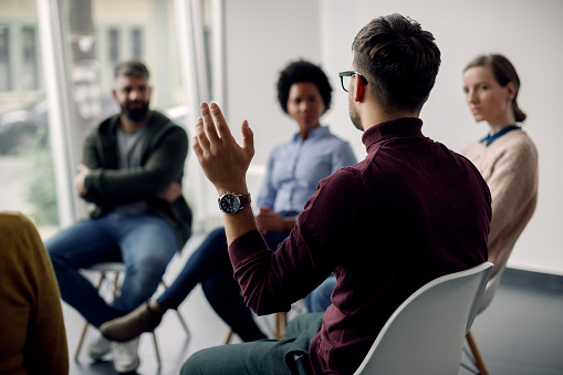 Rear view of man raising his hand to ask a question while attending group therapy.