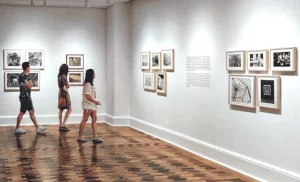 People look around art gallery space with white walls and a wood floor.