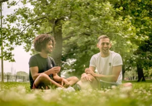 Two men sit in the grass with green trees behind them.