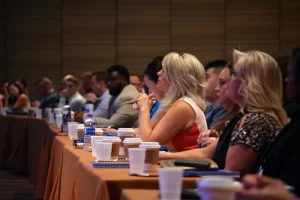 People sit at conference table with coffees and waters, listening to a speaker.