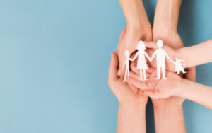 Three pairs of hands placed on top of each other, holding a paper cut-out of a family. 