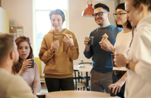 People standing around a table eating snacks and coffee while listening to a speaker.