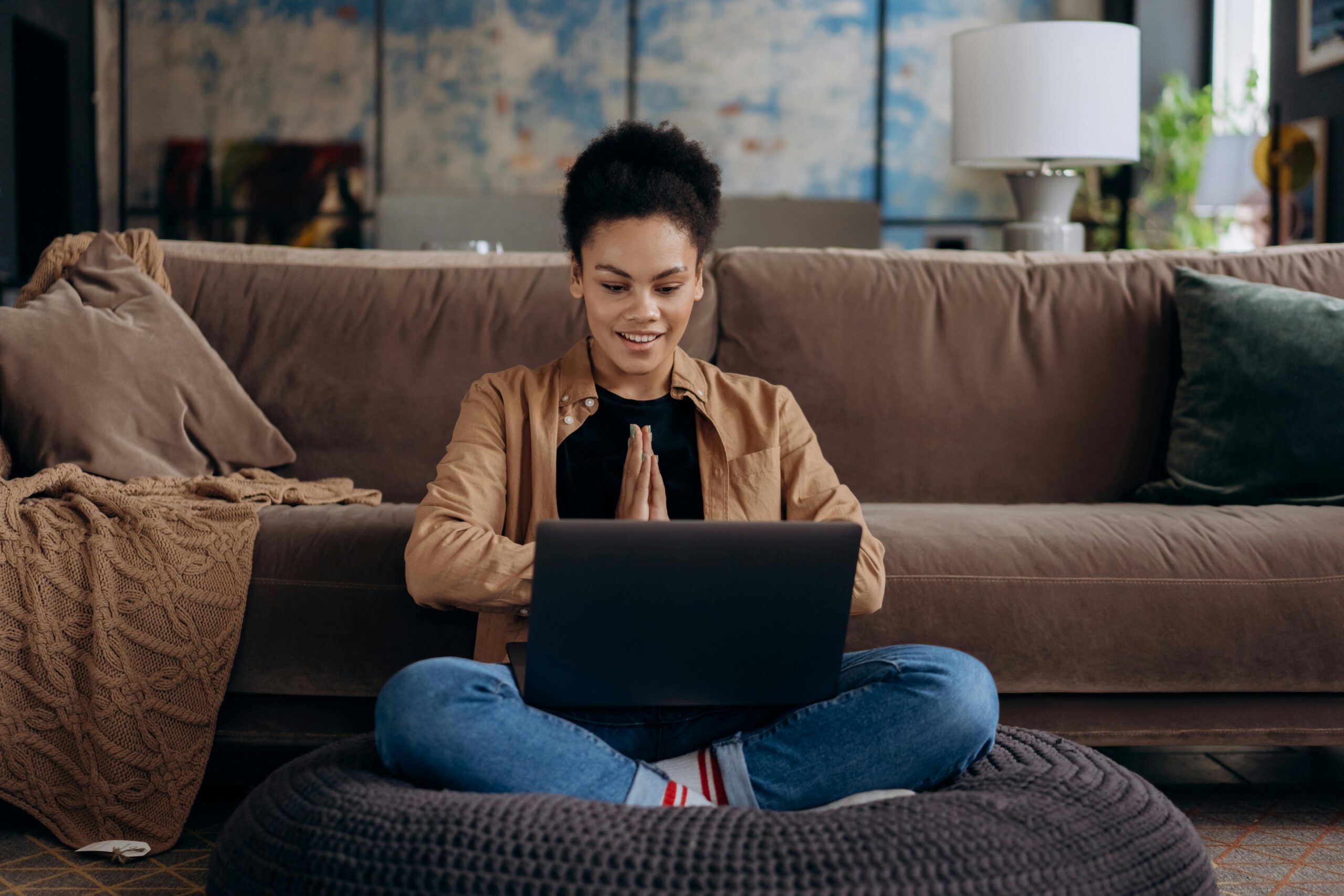Woman sits against a brown couch looking at her laptop