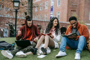 Three students sit on the lawn of a college campus in the fall