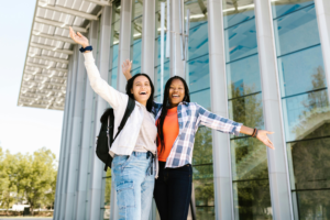 Two college students happily jump in front of a glass building