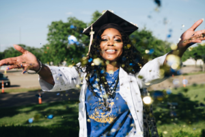 A graduate wearing a graduation cap smiles and throws sparkly blue and gold confetti in front of their face
