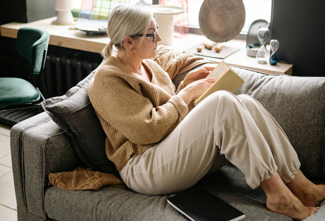 Women sits on a couch in a home, holding a book and looking out the window