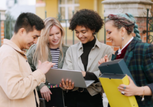 Four students stand together looking at a laptop outdoors