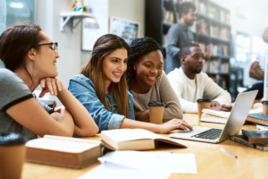 Students sit at a table in a library, smilling and looking at a laptop screen