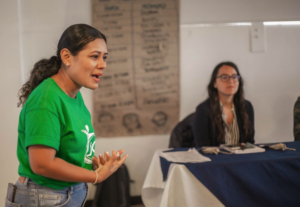One woman, dressed in green, speaks while another woman listens at a table