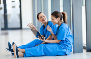 Two nurses dressed in blue scrubs sit in a hallway and laugh while looking at a tablet computer
