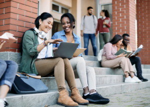 Students sit on concrete steps working on a laptop and smiling