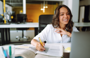 Woman sits in front of laptop smiling and taking notes